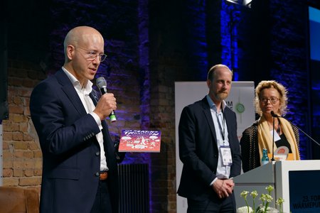Ein Mann steht am Mikrofon und hält ein Buch in der Hand. Hinter ihm befinden sich zwei weitere Personen, die aufmerksam zuhören. Die Szene spielt sich auf einer Bühne während einer Veranstaltung ab, mit beleuchtetem Hintergrund und einer Tafel im Vordergrund.