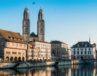 Ein Blick auf die Altstadt von Zürich entlang des Wassers. Im Hintergrund ragen die Türme der Stadtkirche über die umliegenden Gebäude. Die Szene zeigt eine klare, sonnige Tagesstimmung und die Reflexion der Architektur im Wasser.