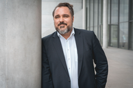 Ein Mann mit Bart trägt einen dunklen Anzug und steht entspannt an einem Säulenstütze. Er hat kurze, leicht gewellte Haare und blickt direkt in die Kamera, wobei er lächelt. Der Hintergrund zeigt eine moderne Architektur.