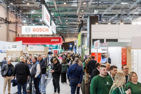 Eine belebte Messehalle mit vielen Besuchern, die zwischen den Ausstellungsständen umhergehen. Im Hintergrund sind Banner von verschiedenen Marken, darunter Bosch und pewo, sichtbar. Die Atmosphäre ist geschäftig und bunt.