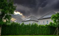 Drei moderne Häuser mit schrägen Dächern und Solarpanelen sind hinter einem hohen grünen Hecken sichtbar. Der Himmel ist dunkel und bedrohlich, mit grauen Wolken und einem schwachen Lichtschein am Horizont.