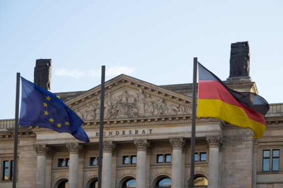 Das Bild zeigt die Flaggen der Europäischen Union und Deutschlands vor dem Gebäude des Bundesrats. Über dem Eingangsbereich ist der Schriftzug „Bundesrat“ sichtbar. Der Himmel ist klar und blau.