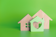 Drei stilisierte Holzhäuser stehen vor einem grünen Hintergrund. Das mittlere Haus ist grün und hat ein Herz-Ausschnitt, während die beiden anderen Häuser aus Naturholz bestehen und unterschiedliche Designs aufweisen.