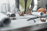 Eine Person in einer gelben Warnweste arbeitet an einem Schreibtisch. Sie skizziert mit einem Stift auf einem großen Bauplan, umgeben von Plänen, einem Lineal und weiteren Werkzeugen. Ein Bauhelm liegt in der Nähe.