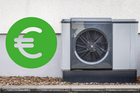 Eine Wärmepumpe steht an einer Wand, daneben ist ein großes grünes Euro-Symbol. Die Pumpe hat ein graues Gehäuse und einen sichtbaren Lüfter. Der Boden ist mit kleinen Steinen bedeckt.