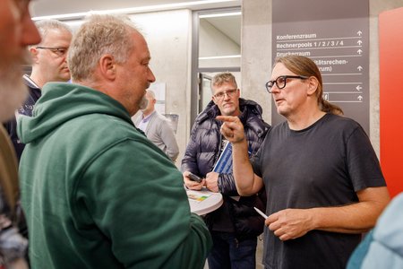 Eine Gruppe von fünf Personen steht in einem Raum. Ein Mann mit Brille und langen Haaren diskutiert intensiv mit einem anderen Mann im grünen Pullover. Die Atmosphäre wirkt angespannt, während die anderen Zuhörer aufmerksam beobachten. Wegweiser zu Konferenzräumen sind im Hintergrund sichtbar.