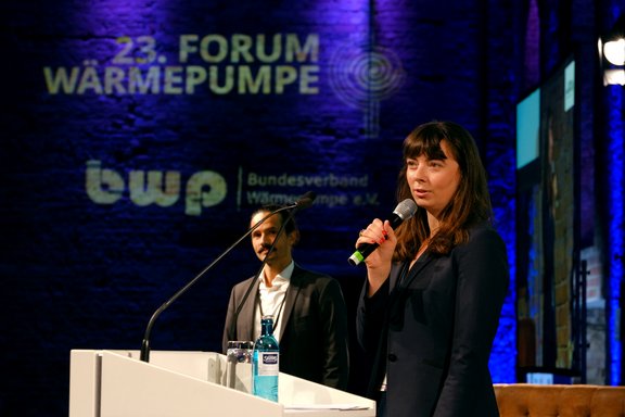 Eine Frau steht am Mikrophon und spricht auf einer Bühne. Im Hintergrund sind Logos und der Schriftzug "23. FORUM WÄRMEPUMPE" zu sehen. Ein Mann steht hinter ihr und hört zu. Die Bühne ist atmosphärisch beleuchtet.