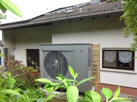 Ein graues Außengerät (vermutlich eine Wärmepumpe) steht neben einem Wohnhaus. Im Hintergrund sind Fenster mit Gardinen und ein Dach mit Solarpanels zu sehen. Um das Gerät wachsen grüne Sträucher und Pflanzen.