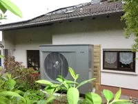 Ein graues Außengerät (vermutlich eine Wärmepumpe) steht neben einem Wohnhaus. Im Hintergrund sind Fenster mit Gardinen und ein Dach mit Solarpanels zu sehen. Um das Gerät wachsen grüne Sträucher und Pflanzen.