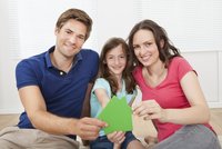Eine Familie sitzt auf dem Boden in einem hellen Raum. Der Vater und die Mutter halten mit der Tochter ein grünes Pappsymbol eines Hauses. Alle lächeln glücklich in die Kamera, was eine positive und familiäre Atmosphäre vermittelt.