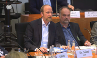 Zwei Männer sitzen an einem Tisch in einem Sitzungssaal. Der Mann links mit Bart spricht, während der andere, mit ernstem Gesichtsausdruck, ihm zuhört. Auf dem Tisch liegen Papiere und Wassergläser. Ein Mikrofon ist sichtbar. Im Hintergrund sieht man verschwommene Silhouetten von weiteren Personen.