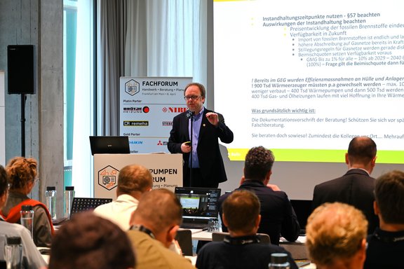 Ein Referent steht vor einem Publikum während eines Fachforums. Auf einem Bildschirm im Hintergrund sind Informationen und Daten zu sehen. Teilnehmer hören aufmerksam zu, einige machen Notizen. Die Atmosphäre ist professionell und informativ.