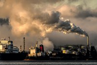 Industrieanlage am Wasser mit Rauch, der aus mehreren Schornsteinen aufsteigt. Im Vordergrund ist ein Schiff zu sehen. Der Himmel ist bewölkt, wobei die Rauchwolken in verschiedenen Grautönen leuchten. Das Bild vermittelt einen Eindruck von Umweltverschmutzung.