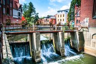 Eine Wasserlauf-Regulierung mit mehreren Ventilen ist die zentrale Darstellung des Bildes. Wasser fließt über die Stufen der Anlage in einen darunterliegenden Kanal. Im Hintergrund sind historische Gebäude und Bäume zu sehen, die eine städtische Umgebung widerspiegeln.