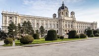 Ein prächtiges historisches Gebäude mit einer beeindruckenden Fassade und vielen Fenstern. Vor dem Gebäude gibt es gepflegte Grünanlagen mit runden Hecken und Nadelbäumen. Der Himmel ist blau und wolkenlos, was eine einladende Atmosphäre schafft.