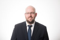 Porträt eines Mannes mit Glatze und Vollbart, der einen dunklen Anzug mit krawatte trägt. Er sieht freundlich aus und steht vor einem weißen Hintergrund.