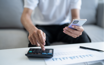 Ein Mensch sitzt auf einem Sofa und hält ein Smartphone in der rechten Hand, während er mit der linken Hand einen Taschenrechner bedient. Vor ihm liegt ein Blatt Papier mit Zahlen und Notizen.