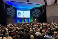 Ein großer Saal ist mit Menschen gefüllt, die auf einer Veranstaltung sitzen. Auf der Bühne stehen zwei Personen, während hinter ihnen eine Präsentation zu sehen ist. Die Atmosphäre ist informativ und die Zuschauer scheinen aufmerksam zuzuhören.