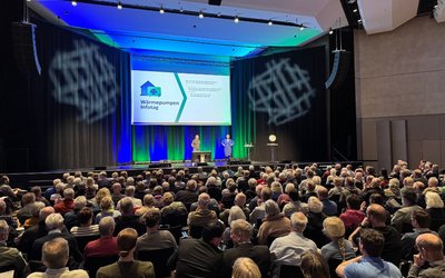 Ein großer Saal ist mit Menschen gefüllt, die auf einer Veranstaltung sitzen. Auf der Bühne stehen zwei Personen, während hinter ihnen eine Präsentation zu sehen ist. Die Atmosphäre ist informativ und die Zuschauer scheinen aufmerksam zuzuhören.