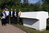 Eine Gruppe von sechs Personen steht auf einem gepflasterten Weg vor einer großen, modernen weißen Struktur. Sie halten Broschüren in den Händen und lächeln. Im Hintergrund sind Bäume und Sträucher sichtbar. Es ist ein sonniger Tag.