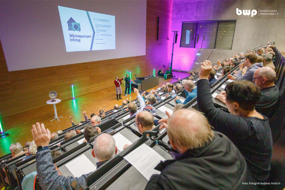 Eine Veranstaltung in einem großen Saal mit zahlreichen Teilnehmenden. Auf der Bühne stehen Personen, die Informationen zu einem Thema präsentieren. Viele Zuhörer heben die Hände, um Fragen zu stellen oder ihre Zustimmung zu zeigen. Im Hintergrund ist eine Präsentationsfolie sichtbar.