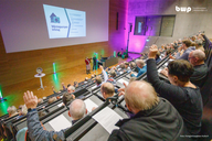 Eine Veranstaltung in einem großen Saal mit zahlreichen Teilnehmenden. Auf der Bühne stehen Personen, die Informationen zu einem Thema präsentieren. Viele Zuhörer heben die Hände, um Fragen zu stellen oder ihre Zustimmung zu zeigen. Im Hintergrund ist eine Präsentationsfolie sichtbar.