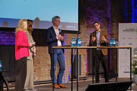 Auf einer Bühne diskutieren vier Personen über aktuelle Themen. Zwei Personen stehen am Tisch und sprechen, während zwei weitere zuhören. Auf dem Tisch stehen Flaschen Wasser. Im Hintergrund ist eine Wand aus Ziegelsteinen zu sehen.