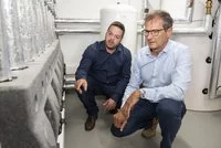 Zwei Männer knien in einem Technikraum. Einer zeigt auf eine technische Vorrichtung, während der andere aufmerksam zuhört. Im Hintergrund sind Rohrleitungen und ein Wasserbehälter sichtbar. Beide Männer tragen formelle Kleidung und scheinen eine fachliche Diskussion zu führen.