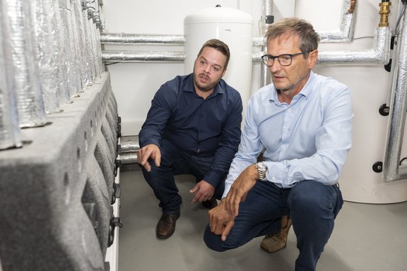 Zwei Männer knien in einem Technikraum. Einer zeigt auf eine technische Vorrichtung, während der andere aufmerksam zuhört. Im Hintergrund sind Rohrleitungen und ein Wasserbehälter sichtbar. Beide Männer tragen formelle Kleidung und scheinen eine fachliche Diskussion zu führen.