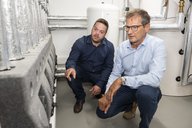 Zwei Männer knien in einem Technikraum. Einer zeigt auf eine technische Vorrichtung, während der andere aufmerksam zuhört. Im Hintergrund sind Rohrleitungen und ein Wasserbehälter sichtbar. Beide Männer tragen formelle Kleidung und scheinen eine fachliche Diskussion zu führen.