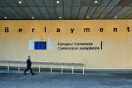 Der Eingang des Berlaymont-Gebäudes mit dem Logo der Europäischen Kommission. Eine Person geht vorbei. Der Schriftzug ist auf mehreren Sprachen zu sehen: „Europese Commissie“ und „Commission européenne“.