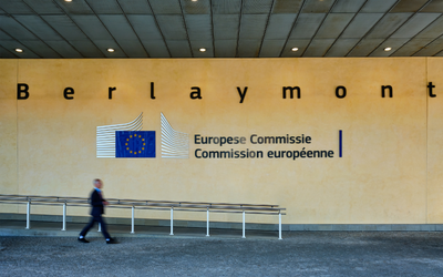 Der Eingang des Berlaymont-Gebäudes mit dem Logo der Europäischen Kommission. Eine Person geht vorbei. Der Schriftzug ist auf mehreren Sprachen zu sehen: „Europese Commissie“ und „Commission européenne“.