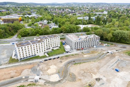 Luftaufnahme einer Baustelle mit zwei Gebäuden: Links ein bereits fertiggestelltes, mehrstöckiges Wohnhaus, rechts ein im Bau befindliches Gebäude. Umgeben von grüner Landschaft und einer Stadtansicht im Hintergrund. Ein unbefestigtes Gelände mit Baustellenmaterial ist im Vordergrund sichtbar.