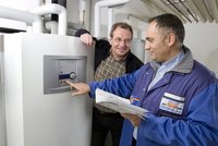 Ein Techniker erklärt einem lächelnden Kunden die Bedienung eines Heizungsgeräts. Der Techniker zeigt auf ein digitales Bedienfeld und hält eine Bedienungsanleitung in der Hand. Die beiden stehen in einem Raum mit Heizungsrohren im Hintergrund.