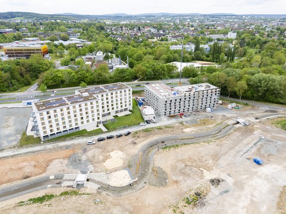 Luftaufnahme eines Baugebiets mit zwei Mehrfamilienhäusern. Das linken Gebäude ist fast fertiggestellt, während das rechte im Bau ist. Umgeben von Grünflächen und einer Straße im Hintergrund. Baustellenfahrzeuge sind sichtbar, ebenso wie unvollendete Erdarbeiten.