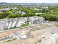 Luftaufnahme eines Baugebiets mit zwei Mehrfamilienhäusern. Das linken Gebäude ist fast fertiggestellt, während das rechte im Bau ist. Umgeben von Grünflächen und einer Straße im Hintergrund. Baustellenfahrzeuge sind sichtbar, ebenso wie unvollendete Erdarbeiten.