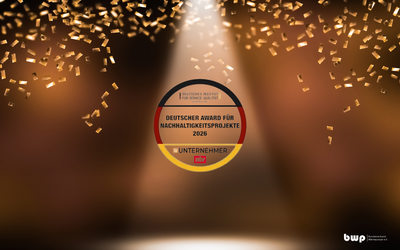 Ein runder Preis mit der Aufschrift „Deutscher Award für Nachhaltigkeitsprojekte 2026“ und dem Wort „Unternehmer“ in der Mitte. Der Hintergrund zeigt eine Bühnenbeleuchtung mit goldenen Konfetti, die eine festliche Atmosphäre schaffen.