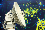 Ein großer Satellitenempfänger steht im Vordergrund, während im Hintergrund ein Radarbild mit gelben und blauen Punkten auf grünem Grund zu sehen ist. Die Grafik zeigt verschiedene Objekte oder Bewegungen, die vom Radar erfasst werden.