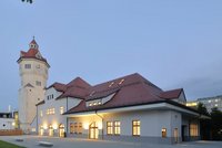 Ein historisches Gebäude mit einem hohen Turm und einem roten Dach, umgeben von sanften Lichtern, die die Fassade beleuchten. Der Himmel ist in der Dämmerung, was eine ruhige Atmosphäre schafft.