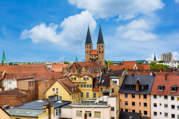 Eine städtische Landschaft mit historischen Gebäuden und zwei markanten Türmen einer Kirche im Hintergrund. Im Vordergrund sind verschiedene Wohnhäuser und Dächer sichtbar. Der Himmel ist blau mit einigen Wolken.