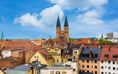 Eine städtische Landschaft mit historischen Gebäuden und zwei markanten Türmen einer Kirche im Hintergrund. Im Vordergrund sind verschiedene Wohnhäuser und Dächer sichtbar. Der Himmel ist blau mit einigen Wolken.