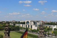 Blick auf das Bundeskanzleramt in Berlin, umgeben von grünen Bäumen und einer sauberen Außenansicht. Im Hintergrund sind verschiedene Gebäudeschornsteine und Kirchtürme sichtbar, bei klarem Himmel mit einigen Wolken.