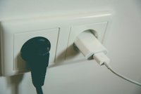 Ein schwarzes und ein weißes Kabel sind in zwei Steckdosen gesteckt. Das schwarze Kabel gehört zu einem Gerät, während das weiße Kabel ein Ladegerät mit einem USB-Anschluss ist. Der Hintergrund ist hell und neutral gehalten.