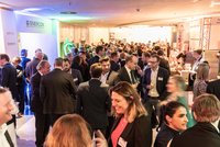Eine belebte Networking-Veranstaltung mit zahlreichen Anwesenden, die in Gruppen miteinander sprechen. Einige Personen halten Getränke in der Hand, während im Hintergrund ein Buffet aufgebaut ist. Die Atmosphäre ist freundlich und geschäftlich, mit bunten Lichtern, die den Raum erhellen.