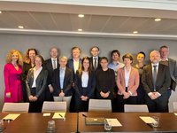 Eine Gruppe von 14 Personen steht vor einem neutralen Hintergrund in einem Konferenzraum. Die Anwesenden sind in formeller Bekleidung und lächeln. Auf dem Tisch sind Schreibmaterialien und Gläser zu sehen. Die Szene vermittelt den Eindruck eines professionellen Treffens oder einer Konferenz.