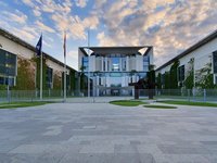 Das Bild zeigt ein modernes Gebäude mit Glasfronten und einem markanten Dach. Vor dem Gebäude stehen Fahnen. Der Platz ist gepflastert und von einem gepflegten Rasen umgeben. Es ist eine leicht bewölkte Himmelsperspektive sichtbar.
