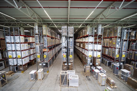 Blick auf ein Lagersystem mit hohen Regalen, in denen verschiedene Produkte verpackt aufbewahrt werden. In der Mitte verläuft ein breiter Gang zwischen den Regalen, die nummeriert sind. Kartons und Geräte stehen auf Paletten.