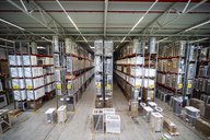 Blick auf ein Lagersystem mit hohen Regalen, in denen verschiedene Produkte verpackt aufbewahrt werden. In der Mitte verläuft ein breiter Gang zwischen den Regalen, die nummeriert sind. Kartons und Geräte stehen auf Paletten.