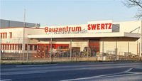Das Bild zeigt ein Bauzentrum mit dem Schriftzug „Bauzentrum SWERTZ“ an der Fassade. Das Gebäude hat eine hellere Außenfarbe mit roten Akzenten. Vor dem Gebäude sind verschiedene Bauutensilien und Geräte sichtbar, und das Gelände ist von einem Zaun umgeben.