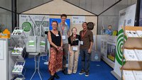 Eine Gruppe von vier Personen steht im Vordergrund eines Messestands. Im Hintergrund sind Informationsmaterialien und Banner zu sehen. Die Personen lächeln und tragen Namensschilder. Die Szenerie vermittelt eine freundliche und einladende Atmosphäre.